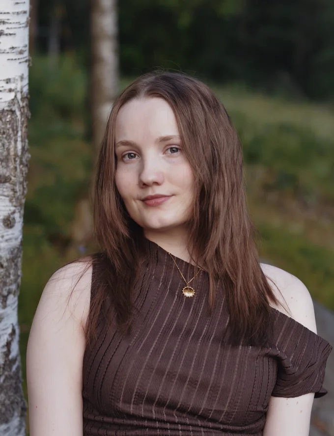 Jenni hymyilee koivun vieressä, kaulassaan kultainen aurinkoriipus